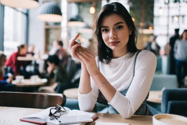 Modern bir kafede boş zamanlarında otururken elleri sımsıkı bağlı, kameraya bakan modaya uygun giyinmiş bir kadın.