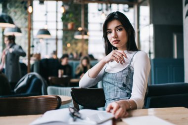 Uzun siyah saçlı, odaklanmış bir kız öğrenci, ara sırasında modern kafedeki ahşap masada otururken kameraya bakıyor.