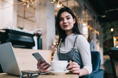 Kafenin bulanık arka planında kahve içerken ve telefondaki mesajları kontrol ederken kameraya bakan genç, gülümseyen bir serbest kadın.