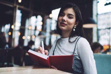 Kafeteryada masada otururken, arka planda bulanık insanlar varken kameraya bakan ve kırmızı düzenleyiciyle notlar alan pozitif kadın serbest yazar.