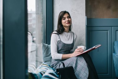 Moda kıyafetleri içinde, kitap okuyan ve rahat bir kafede pencerenin yanında sıcak bir içecek içen genç bir kadının yan görüntüsü.