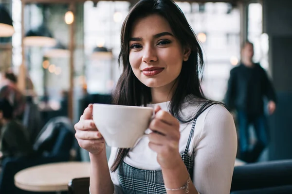身着休闲装 面带微笑的年轻女性坐在灯光咖啡店的模糊背景上 一边看着相机 一边喝着一大杯咖啡 — 图库照片