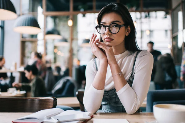 カジュアルな服装でカメラを見ながらノートを手にもたれながらカフェに座っている女子大生は大学の課題に取り組む — ストック写真