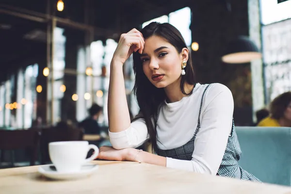 カメラを見ながらコーヒーを片手にテーブルに座り 手にもたれかかる赤い唇のカジュアルな服を着た女性 — ストック写真