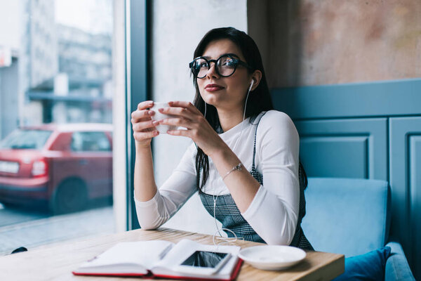 Positive lady in casual wear and eyeglasses looking away and smiling while drinking coffee in cafe and listening to music with earphones