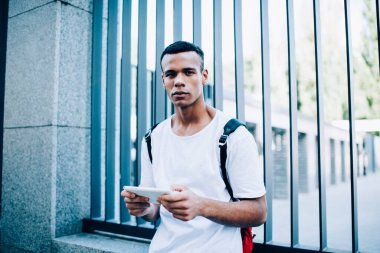 Sırt çantalı dalgın Afro-Amerikan erkek kameraya bakarken cep telefonunda metalin yanında sokak sütunuyla geziyor.