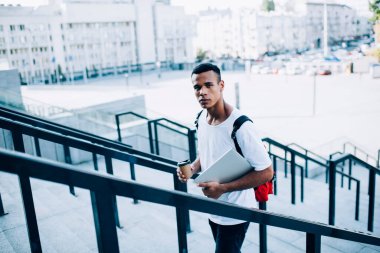 Ciddi, kırmızı sırt çantalı genç bir adam güneşli bir günde metal korkuluklar arasında yürürken kameraya bakıyor.