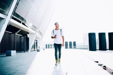 Gündelik kıyafetli, sırtında sırt çantasıyla modern binanın yanında yürüyen ve sıcak içecek içen bir sürü genç adam.