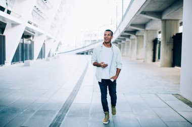 Gündüz vakti şehir caddesinde beyaz bina ve köprü yakınlarında tek başına dolaşırken günlük kıyafetli mutlu siyahi bir adam cep telefonundan mesaj atıyor.