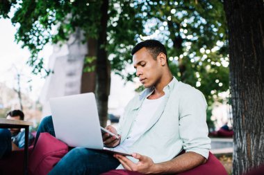 Ciddi genç siyahi bir adam günlük kıyafetleriyle şezlongda oturuyor ve netbook 'ta uzaktan çalışıyor. Bu arada yazın ağaçların yanındaki akıllı telefondan mesaj atıyor.