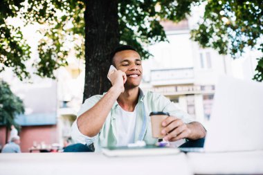 Mutlu siyah erkek serbest çalışan, dişlek gülüşlü ve kağıt bardakta kahveli, caddede oturan ve cep telefonuyla konuşan.