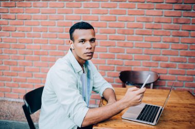 Kendine güveni olan genç siyahi bir adam masanın yanındaki taburede oturup uzaktan internet rehberi üzerinde çalışıyor ve akıllı telefonuyla sokakta kulaklık takıyor ve kameraya bakıyor.