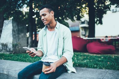 Genç, Afro-Amerikan bir adamın günlük kıyafetleri içinde ve elinde kağıt bardakla parkta oturan cep telefonunun yan görüntüsü.