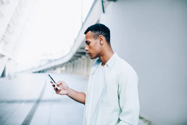 白い橋と昼間の壁の近くの街の通りに一人で立っている間 携帯電話を使用してカジュアルな服装で深刻な若い黒人男性の側面図 — ストック写真