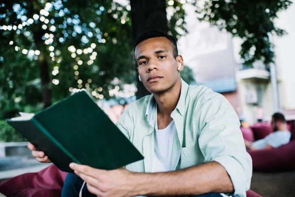 年轻自信的黑人男子穿着衬衫坐在室外休息区 带着一本有趣的书 在白天看着相机 — 图库照片