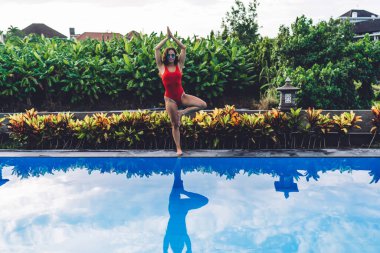 Mayolu ve güneş gözlüklü genç bayan dinlenme yerinin bahçesinde yoga yaparken boş havuzun kenarında, Vrikshasana 'da duruyor.
