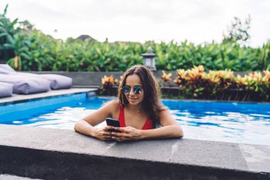 Yuvarlak güneş gözlüklü genç esmer, yüzme havuzunda duruyor ve yaz tatilini egzotik bir otelde geçirirken akıllı telefondan sosyal medyada sörf yapıyor.