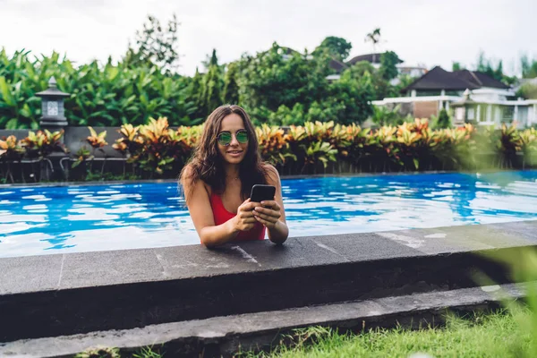 年轻的长发女 戴着时髦的太阳镜 站在游泳池里 用手机在社交媒体上冲浪 在奇异迷人的度假胜地享受暑假 — 图库照片
