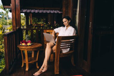 Beyaz tişörtlü, tahta sandalyede çıplak bacaklarıyla oturan ve tropikal villa verandasında serinlerken netbook kullanan tam vücut pozitifliği olan genç esmer.