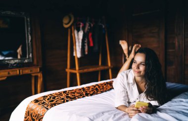 Gündelik giysiler içinde gülümseyen güzel bir bayan güneşli bir yaz gününde akıllı telefon kullanırken başka yere bakıyor ve yatakta uzanıyor.