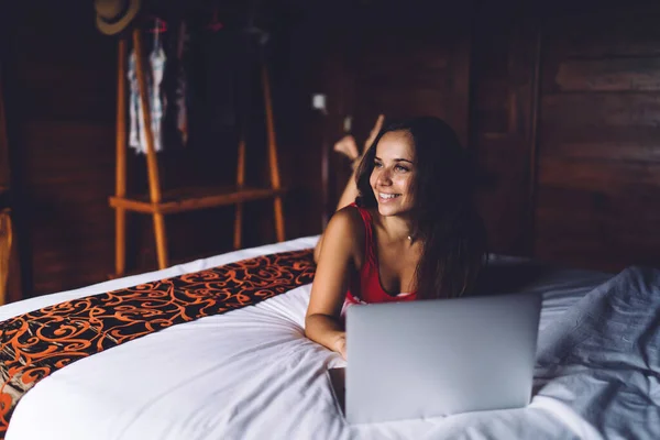 积极的女性躺在舒适的卧室里 通过上网本上网 同时在家里享受周末的快乐时光 并把目光投向远方 — 图库照片