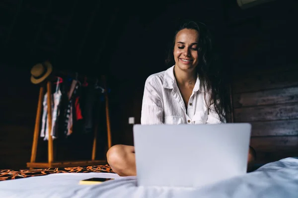 コンテンツスリム女性で白いシャツに座って居心地の良いベッドで足を渡り 光の寝室で時間を過ごしながら現代的なネットブックを使用 — ストック写真