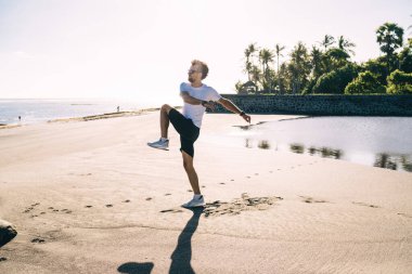 Spor kıyafetleri ve sakallı güneş gözlüklü genç adam sıkı çalışmadan önce boş kumlu sahilde egzersiz yapıyor.