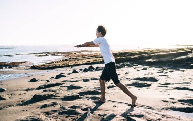 Güneşli bir günde, deniz kenarındaki ıslak kumlu sahilde Warrior pozu veren uzun boylu, spor kıyafetli bir adamın yan görüntüsü.