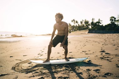 Siyah şortlu sportif beyaz adam, Malibu 'da deniz dalgalarını yakalamak için kumlu sahilde sörf yapma becerileri geliştiriyor. Genç adam deniz kıyısında ısınma antrenmanı yapıyor.