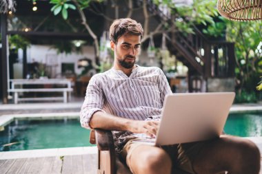 Çizgili tişörtlü mutlu erkek internette geziniyor ve havuz kenarındaki terasta dinlenirken internet üzerinden daktilo yazıyor.