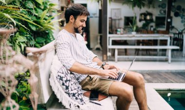 Yan manzara, modaya uygun yaz kıyafetleri içinde neşeli bir erkek modern dizüstü bilgisayarda daktilo kullanıyor ve egzotik bir tatil beldesinde yüzme havuzunun yanındaki bankta dinlenirken gülümsüyor.