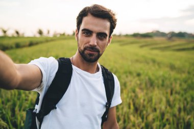 Beyaz tişörtlü yakışıklı genç bir erkek. Elinde kamera var ve yemyeşil bir çayırda durup kameraya bakarken selfie çekiyor.
