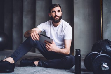 Spor giyim sektöründe sakallı genç bir adam spor salonunda antrenman yaparken tablet kullanıyor ve dinleniyor.