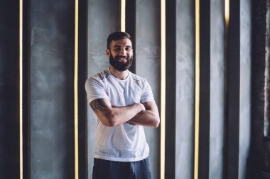 Aktif giyinmiş neşeli sakallı bir erkeğin yarım boy portresi sağlıklı yaşam tarzının keyfini çıkarıyor ve yakışıklı profesyonel sporcu spor salonunda kollarını kavuşturup gülümseyerek kameraya bakıyor.