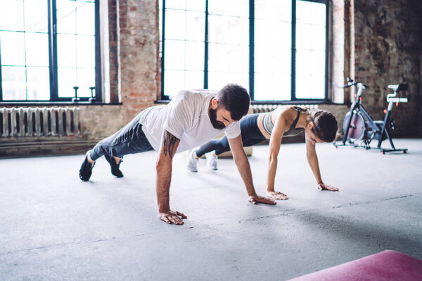 Full length of focused sportswoman in activewear doing plank exercise on straight hands during workout in spacious gym with fitness trainer