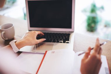 Tahta masada elinde kahve ve not defteriyle oturan ve dizüstü bilgisayarını proje üzerinde çalışmak için kullanan yüzü olmayan kadın serbest yazardan.