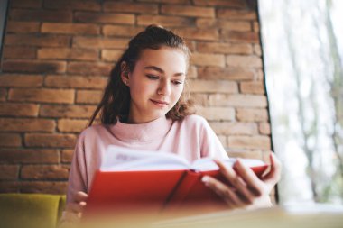 Gündüz vakti rahat bir kafede vakit geçirirken, kırmızı kaplı, günlük kıyafetlerle kitap okuyan düşük açılı bir kadın.