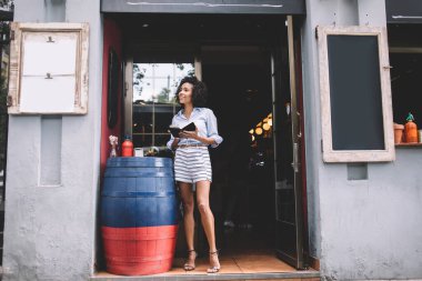 Mavi tişörtlü ve etekli neşeli bayan hostesin elinde defteriyle terastaki modern kafenin kapısının ve fıçısının yanında duruyor.