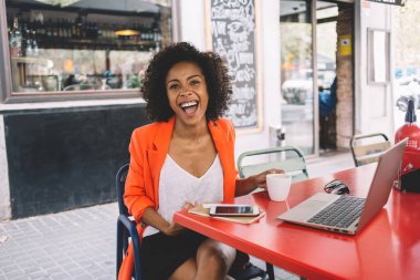 Neşeli genç etnik bayan girişimci bir fincan kahveyle masada oturuyor ve kameraya bakarken dizüstü bilgisayarla çalışıyor.