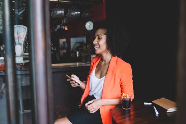 Kafeteryada dinlenirken bir bardak kokteyl ve planlamacıyla masada oturan şık giyinmiş Afro-Amerikan kadının yan görüntüsü.