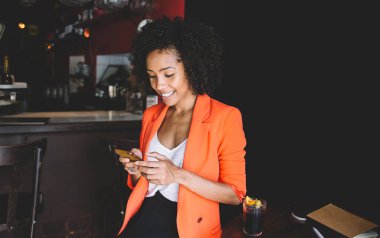 İyimser Afro-Amerikan bayan yönetici şık kıyafetler içinde masada oturmuş, bir bardak içkiyle planlamacı akıllı telefonuyla sohbet ederken rahat bir kafede dinleniyor.
