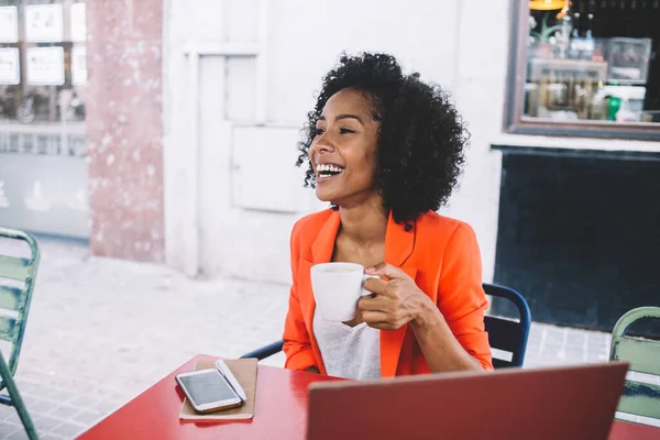 アフリカ系アメリカ人のカジュアルな女性で カフェテリアでノートパソコンの前に座ってお茶を飲みながら笑って離れて見ています — ストック写真