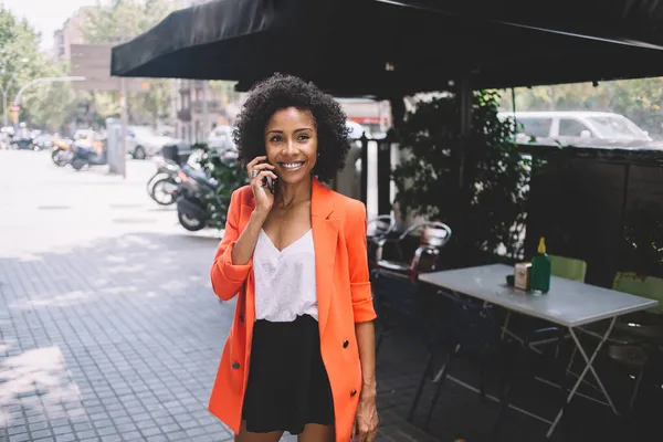 ストリートカフェの近くの通りに立っている間 流行の鮮やかな衣装で笑顔とカメラを見て幸せな若い黒人女性 — ストック写真