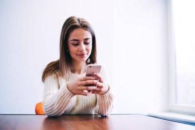 Uzun saçlı bir kadın aydınlık odadaki ahşap masada oturuyor ve sosyal ağdaki mesajları kontrol etmek için akıllı telefonu kullanıyor.