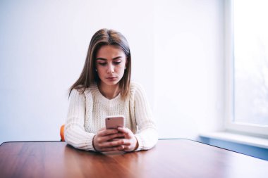 Beyaz kazaklı, uzun saçlı, düşünceli bir kadın pencerenin yanındaki tahta masada oturuyor ve kırık telefonuyla sörf yapıyor.