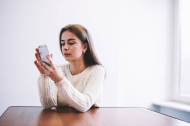 Uzun saçlı, sıradan giysili genç bir kadın masada oturuyor ve mesaj atmak için modern cep telefonunu kullanıyor.