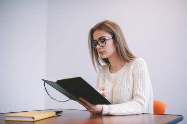 Düşünceli bayan öğrenci, günlük giysiler ve gözlükler takmış masaya oturmuş sınav hazırlıkları sırasında not defterinde notlar okuyordu.