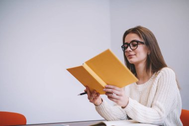 Gündelik kıyafetli genç kız öğrenci aydınlık odada oturuyor ve yeni bilgiler öğrenirken ders kitabı okuyor.