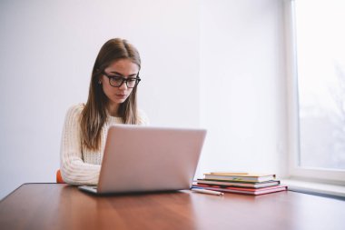 Gözlüklü ve beyaz kazaklı düşünceli genç bayan planlamacı ve kalemle pencerenin yanında otururken çağdaş netbook 'a göz atıyor.