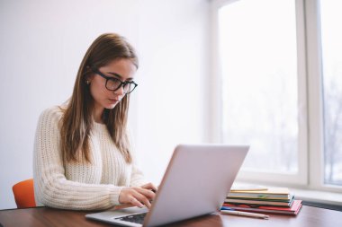 Gözlüklü ve beyaz kazaklı meşgul genç bir kadın, hafif çalışma alanındaki ahşap masada çalışırken taşınabilir laptopta bilgileri kontrol ediyor.